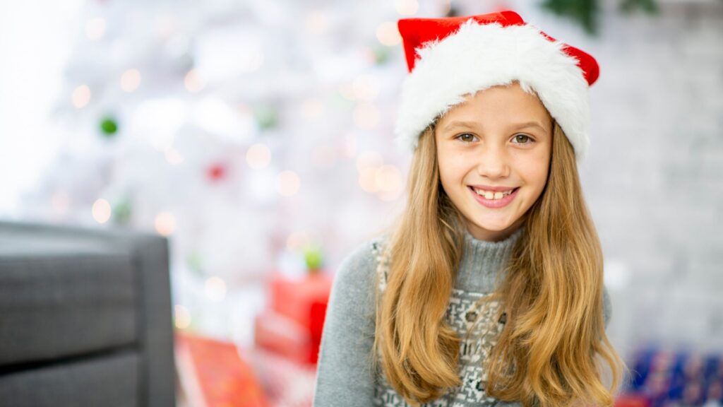 girl in a santa hat sits in front of christmas tree to hear stocking stuffer ideas