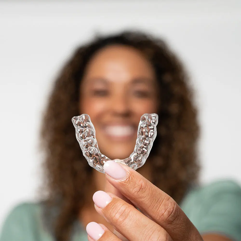 Young woman holding up clear aligner, smiling in the background, San Antonio orthodontist