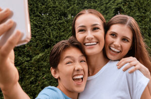 Mother, daughter and son smiling looking at phone with green hedge background, braces and clear aligners at Senties Family Orthodontics in Alamo Ranch, San Antonio